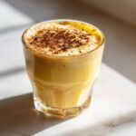 Close-up of a glass filled with creamy, yellow eggnog, topped with foamed milk and a dusting of nutmeg.
