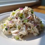 A mound of creamy greek yogurt chicken salad mixed with celery and red onion served on a white plate.