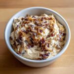A close-up of creamy onion dip topped generously with caramelized or fried onions in a white bowl.