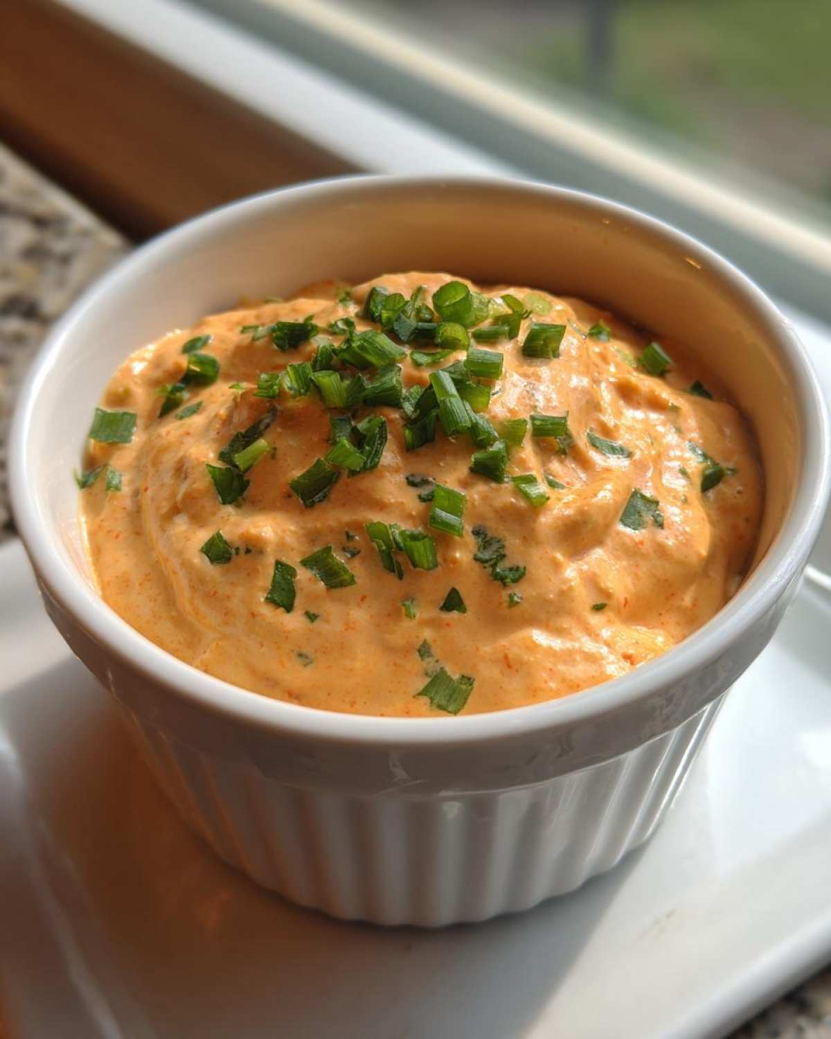 Close-up of a small white ramekin filled with orange-hued remoulade sauce, garnished heavily with chopped green chives.