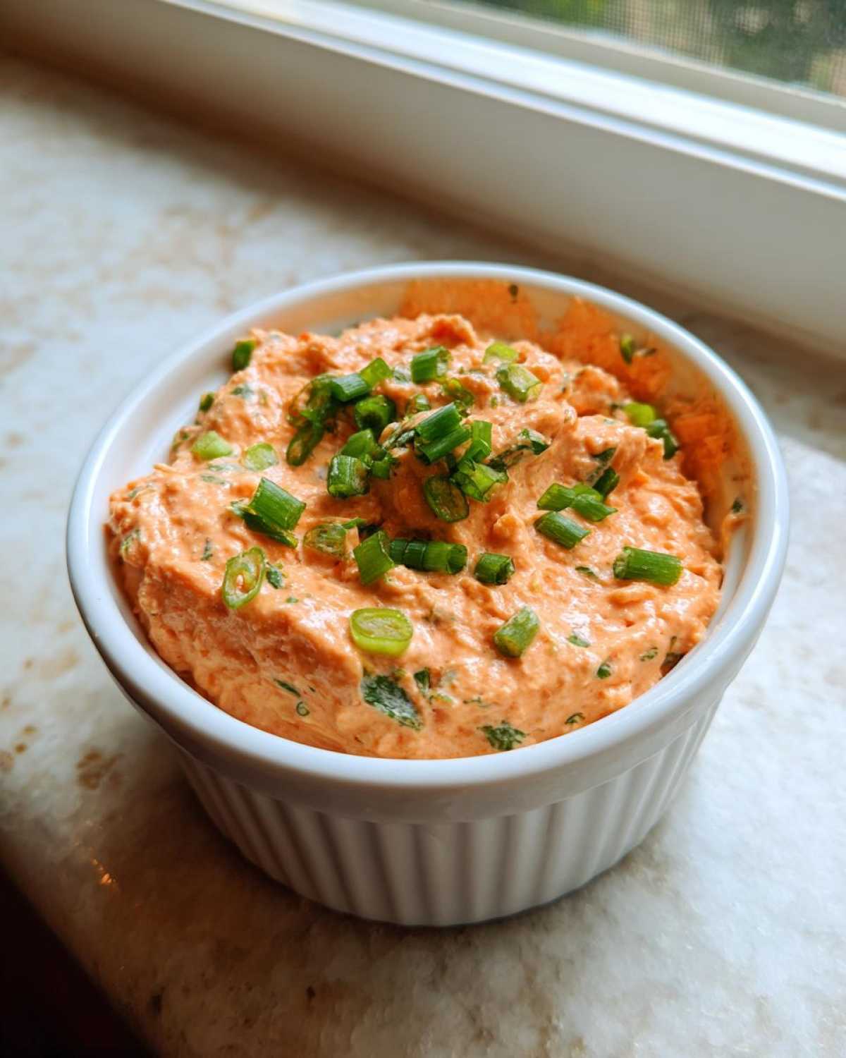 A small white ramekin filled with creamy, orange-tinted remoulade sauce, garnished generously with chopped green onions.