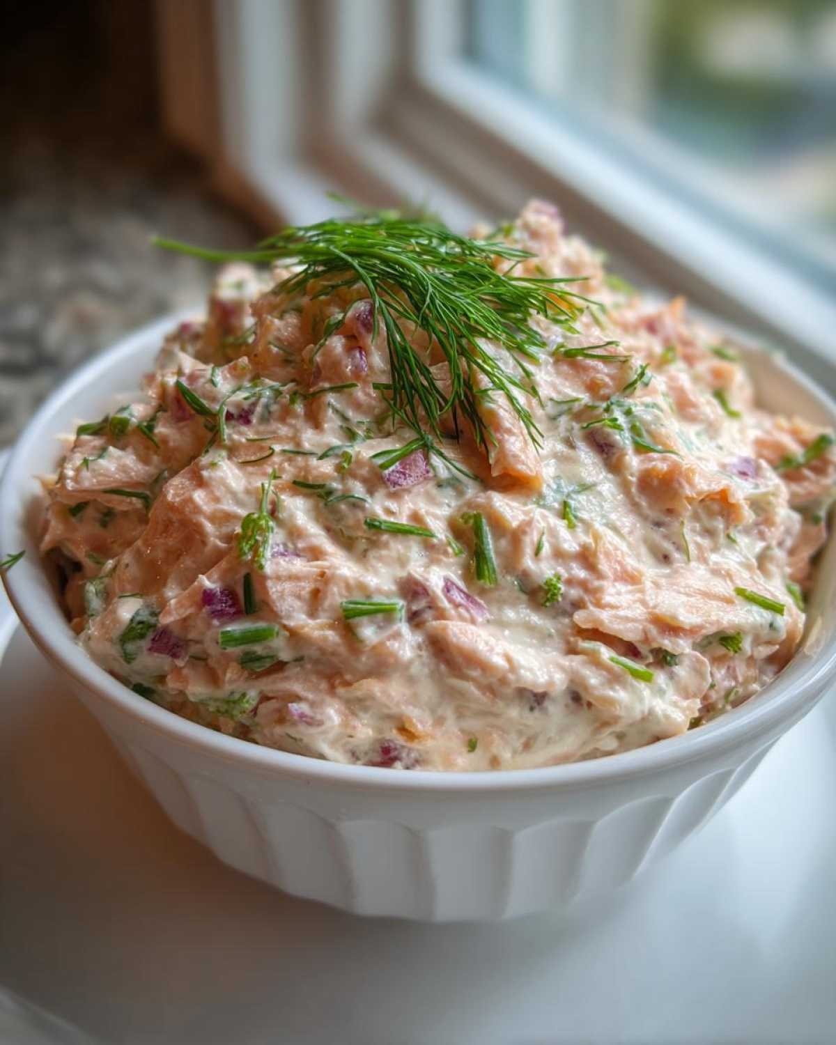 Close-up of creamy smoked salmon dip mixed with red onion and chives, topped with fresh dill.
