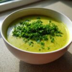 A close-up of vibrant green zucchini soup topped generously with chopped fresh parsley in a white bowl.