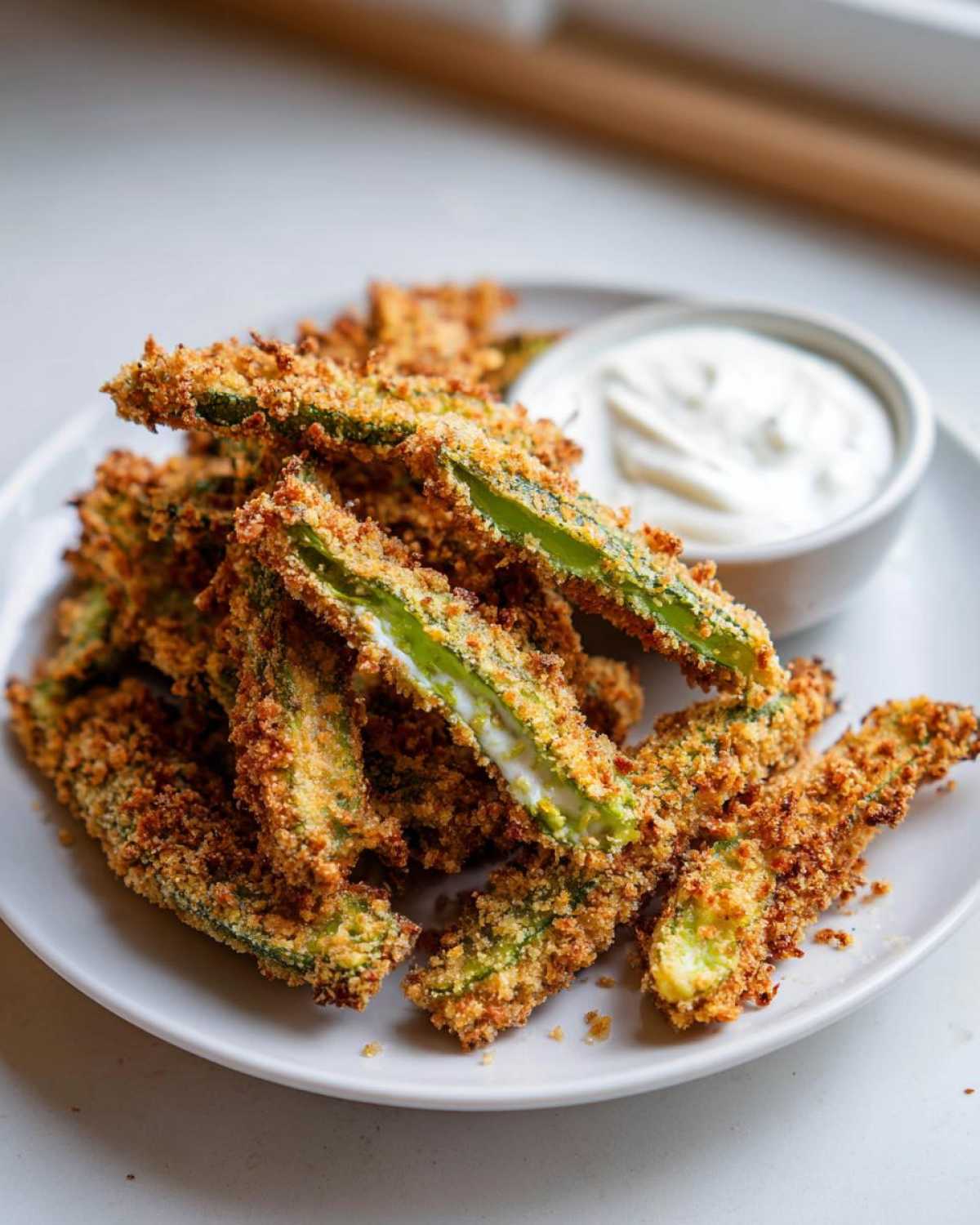A pile of golden brown, crispy Fried Pickles served on a white plate next to a small bowl of white dipping sauce.