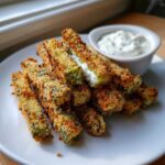 A pile of golden brown, crispy Fried Pickles served on a white plate next to a small bowl of white dipping sauce.