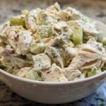 A close-up view of creamy dill pickle chicken salad, featuring chunks of chicken and cucumber, served in a white bowl.