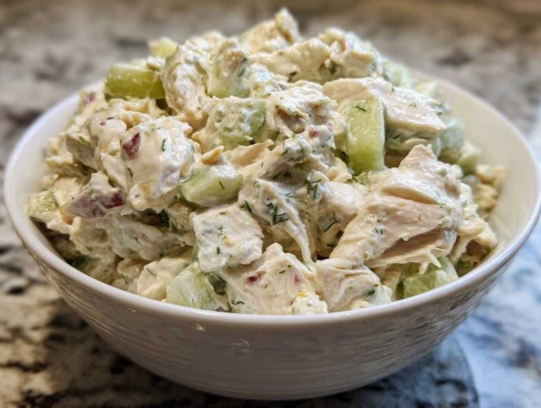 A close-up view of creamy dill pickle chicken salad, featuring chunks of chicken and cucumber, served in a white bowl.