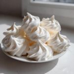 A pile of perfectly piped, bright white meringue cookies displayed on a small white plate near a window.