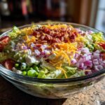 Close-up of a vibrant 7 layer salad in a clear glass bowl, topped with bacon, cheese, and red onion.