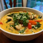 A close-up of a white bowl filled with rich, yellow coconut curry soup, topped with chicken, red peppers, spinach, and fresh cilantro.