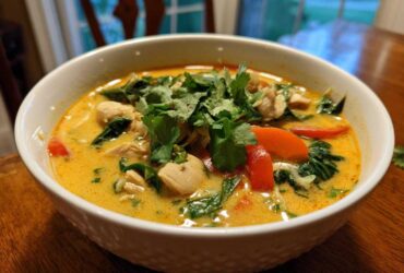A close-up of a white bowl filled with rich, yellow coconut curry soup, topped with chicken, red peppers, spinach, and fresh cilantro.