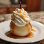 A single, glistening poached pears topped with a swirl of whipped cream and drizzled with caramel sauce on a white plate.