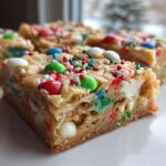 A close-up of a thick, chewy slice of Christmas cookie bars topped with colorful candies and sprinkles.