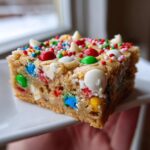 A thick square of chewy christmas cookie bars topped with colorful candies and white chocolate chips.