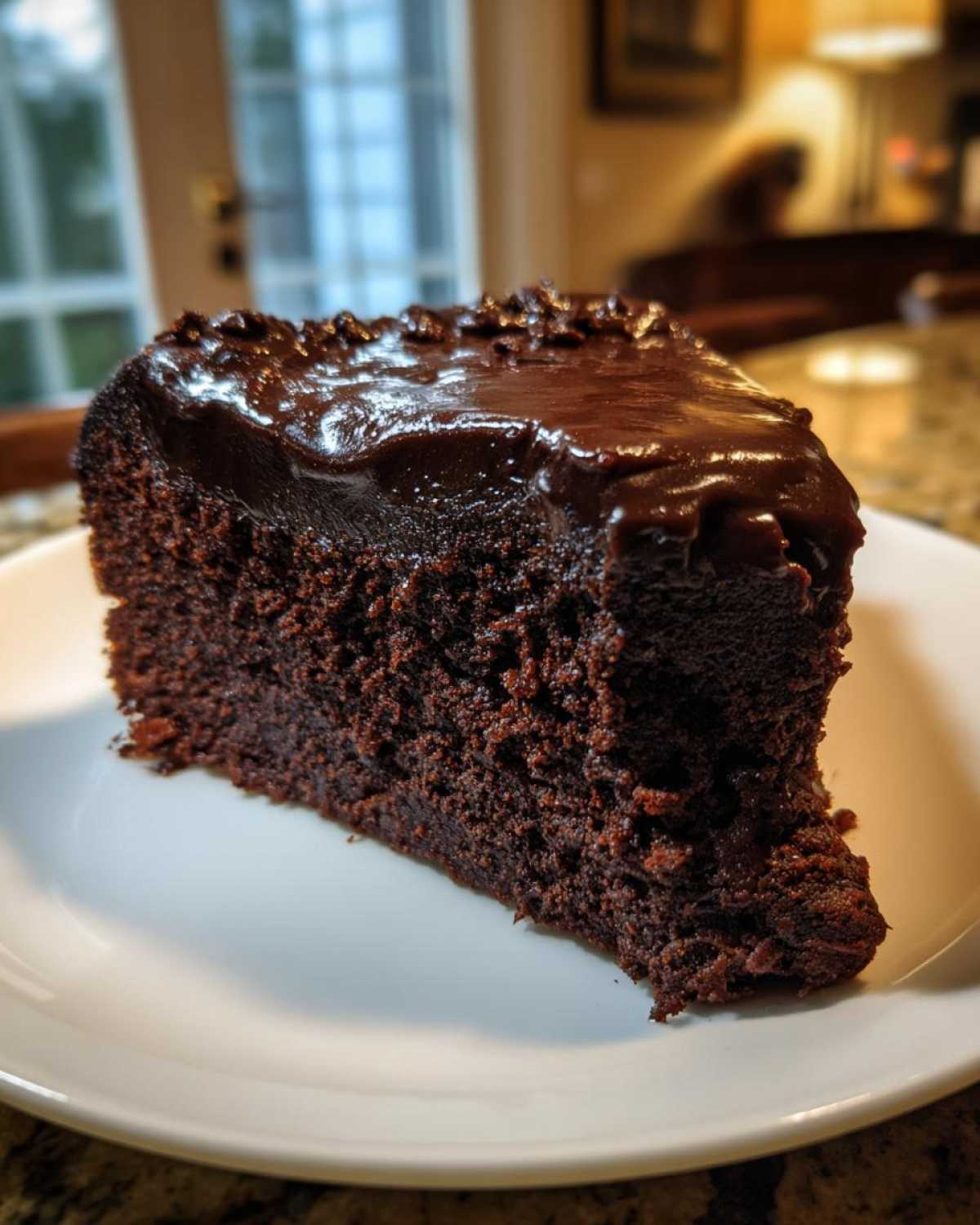 A rich, dark slice of flourless chocolate cake topped with shiny chocolate ganache, served on a white plate.