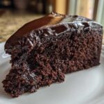 A rich, moist slice of flourless chocolate cake topped with shiny chocolate ganache, served on a white plate.