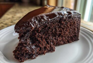 A rich, moist slice of flourless chocolate cake topped with shiny chocolate ganache, served on a white plate.