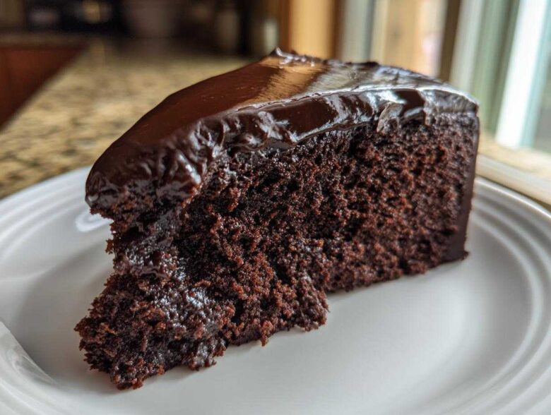 A rich, moist slice of flourless chocolate cake topped with shiny chocolate ganache, served on a white plate.