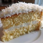 Close-up of a moist slice of coconut cake showing layers of yellow sponge, white frosting, and topped with shredded coconut.