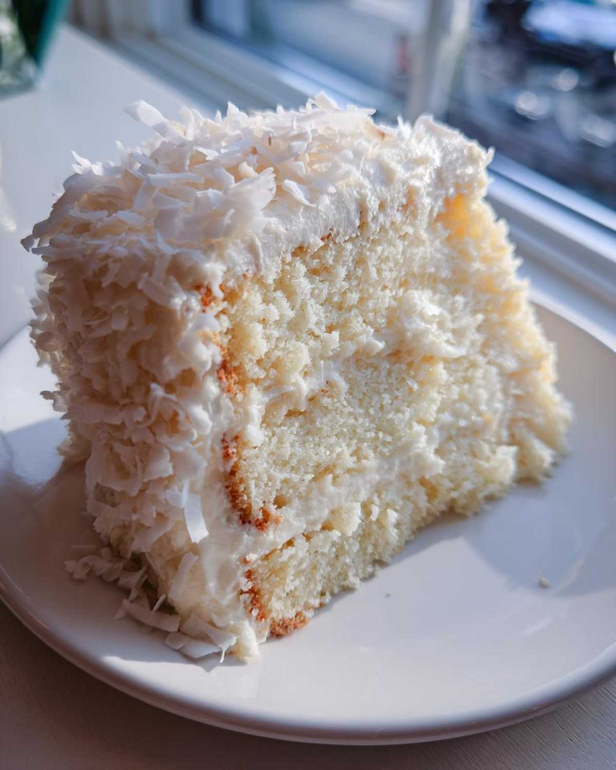 A close-up slice of fluffy coconut cake covered in white frosting and shredded coconut flakes.