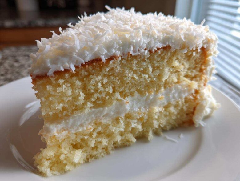 Close-up of a moist slice of coconut cake showing layers of yellow sponge, white frosting, and topped with shredded coconut.