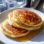 A close-up stack of three golden brown, fluffy pancakes drizzled with maple syrup.