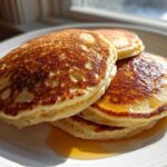 A close-up stack of three golden brown, fluffy pancakes drizzled with maple syrup on a white plate.