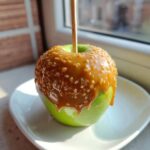 A bright green apple covered in thick, dripping caramel and chopped nuts, ready to eat as a caramel apple.