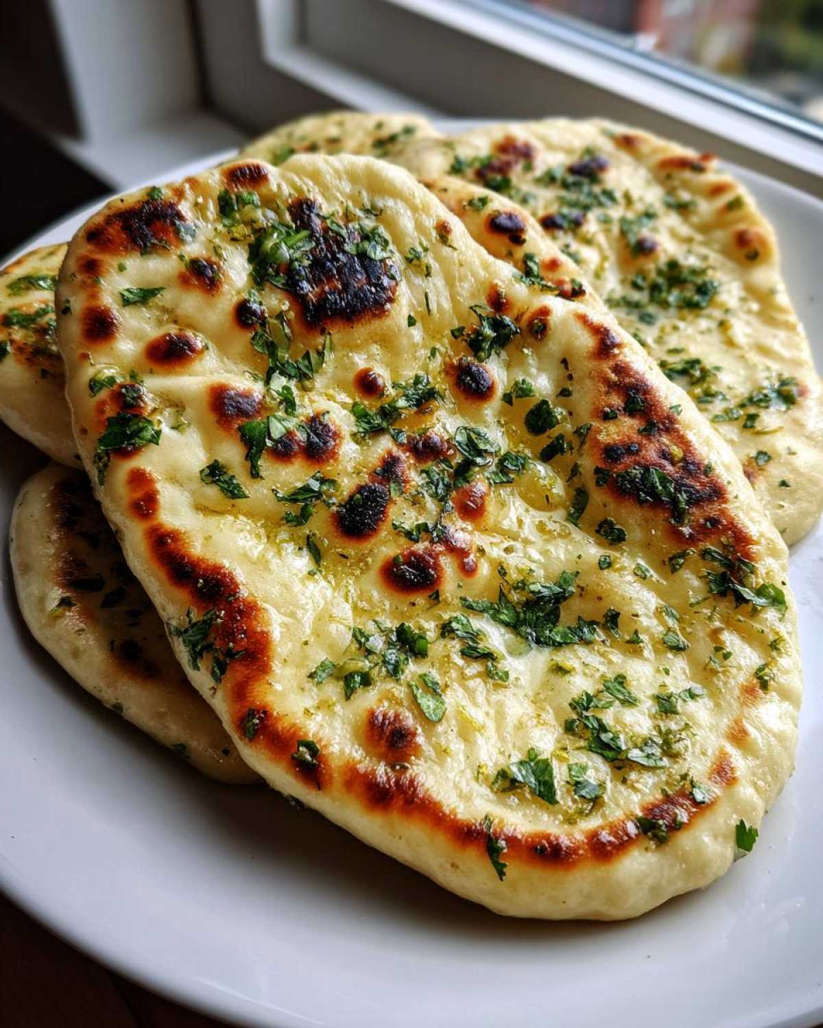 Close-up of several pieces of soft, charred naan bread brushed with butter and topped with fresh green herbs.
