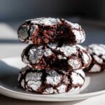 A stack of three fudgy chocolate crinkle cookies dusted with powdered sugar, one cookie on top has a bite taken out.