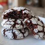 A stack of four rich, dark chocolate crinkle cookies dusted heavily with powdered sugar, one cookie broken open to show the fudgy interior.