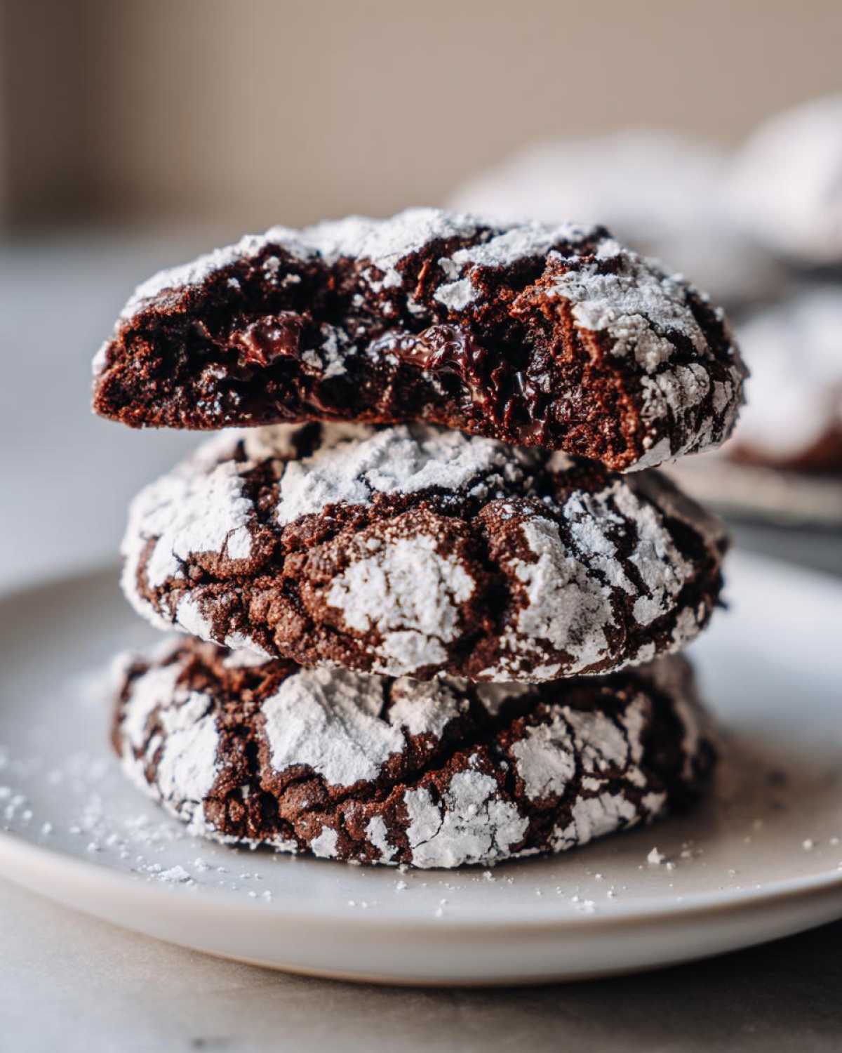 A stack of three fudgy chocolate crinkle cookies coated in cracked white powdered sugar, resting on a white plate.