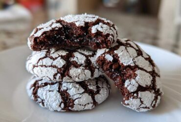 A stack of four rich, dark chocolate crinkle cookies dusted heavily with powdered sugar, one cookie broken open to show the fudgy interior.