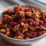 Close-up of a light gray bowl filled with rich, spiced gingerbread granola mixed with bright red dried cranberries.