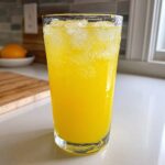 A tall glass filled with bright yellow, fizzy lemonade and crushed ice cubes, sitting on a white countertop.
