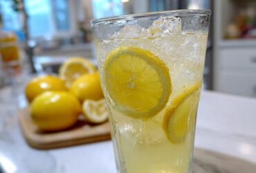 Close-up of a tall glass filled with bubbly, ice-cold lemonade and lemon slices, with whole lemons blurred in the background.