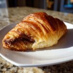 A freshly baked, golden brown chocolate croissant showing rich, flaky layers and melted chocolate filling.