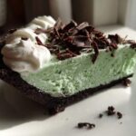 Close-up of a slice of bright green grasshopper pie with a dark chocolate crust, topped with whipped cream and chocolate shavings.