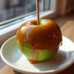 A single green apple coated in thick, dripping caramel, served on a small white plate.