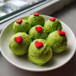 Six bright green Grinch Oreo balls topped with small red heart sprinkles on a white plate.