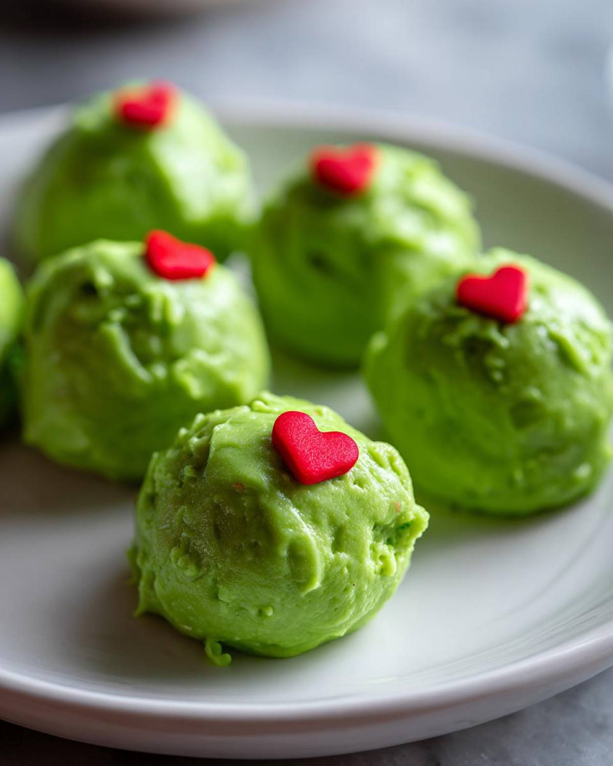 Close-up of bright green Grinch Oreo balls topped with small red candy hearts on a white plate.