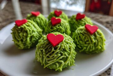 Six festive grinch oreo balls covered in green frosting strands and topped with small red hearts.