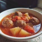 Close-up of a white bowl filled with rich, tomato-based albondigas soup featuring meatballs, potatoes, and carrots.