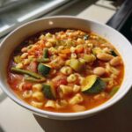 Close-up of a white bowl filled with rich minestrone soup featuring zucchini, beans, carrots, and small pasta shapes.