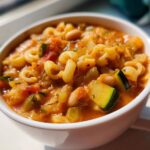 A close-up shot of a white bowl filled with rich, tomato-based minestrone soup featuring pasta, zucchini chunks, and white beans.