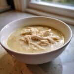 A white bowl filled with creamy, rich cream of chicken soup topped generously with shredded white meat chicken.