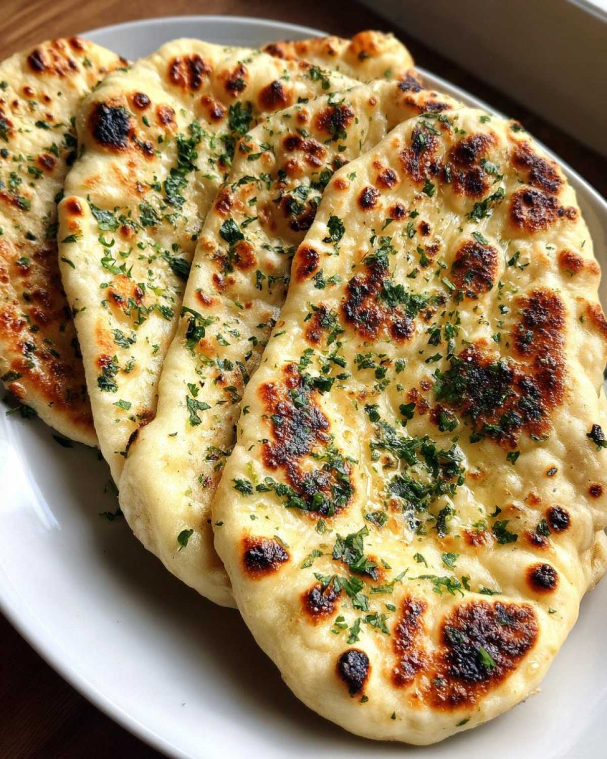 Close-up of three pieces of freshly baked naan bread, charred slightly and topped with melted butter and fresh parsley.