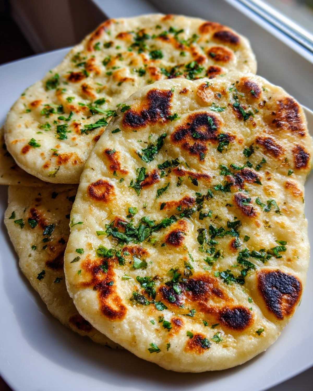 Close-up of three pieces of freshly cooked naan bread, golden brown with charred spots and sprinkled with fresh green herbs.