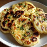 Close-up of several golden, slightly charred pieces of homemade naan bread topped with fresh green herbs.