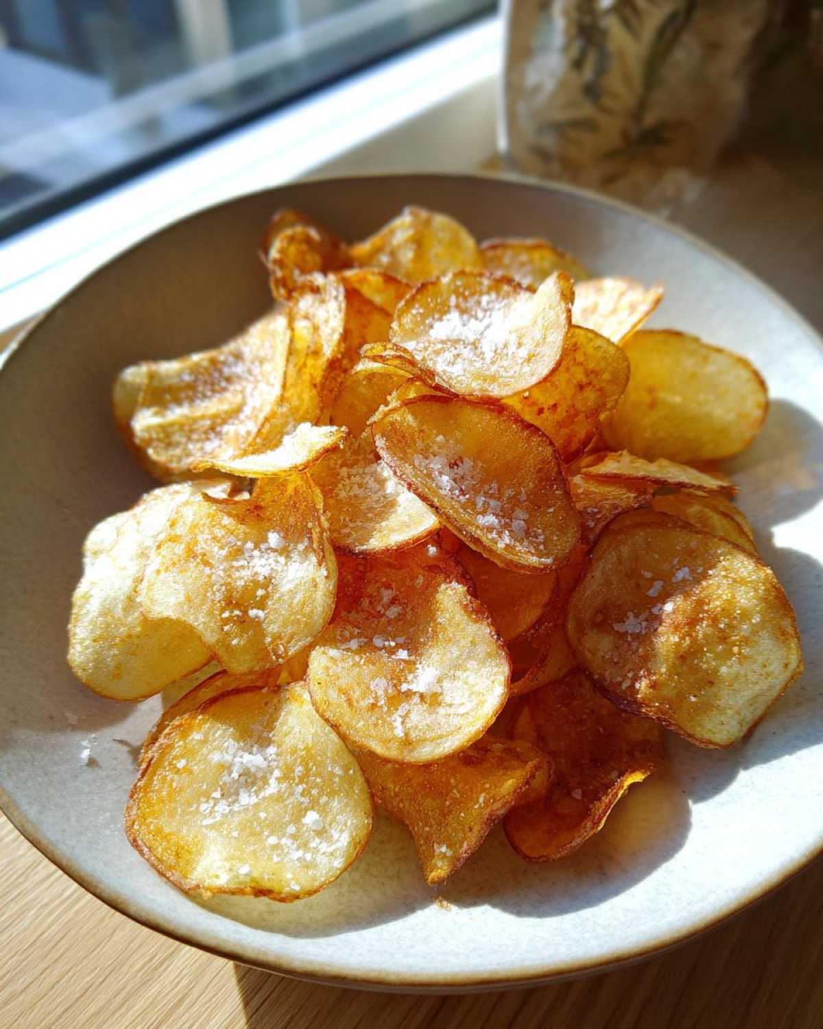 A close-up of homemade, golden brown potato chips generously sprinkled with coarse sea salt in a light-colored bowl.
